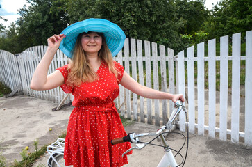 Cheerful woman in a red polka dot dress and a large blue hat stands beside a bicycle near a white wooden fence in a sunny outdoor setting, conveying style and carefree leisure