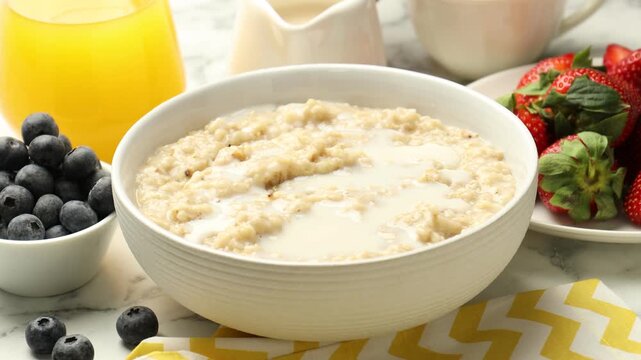 Healthy breakfast. Woman adding milk and berries to oatmeal at white marble table, closeup