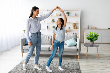 Fototapeta premium Cheerful asian mother and daughter dancing to music, holding hands and enjoying spending time together, having fun in living room interior at home. Full length shot