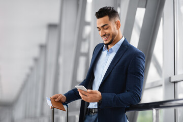 Mobile Roaming. Young Smiling Middle Eastern Businessman Using Smartphone In Airport Terminal,...