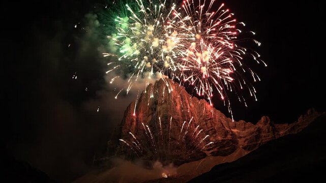Spectacular fireworks burst in emerald and ruby colors above a majestic mountain peak at night, creating a vibrant and celebratory display.