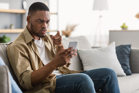 Problem With Cellphone. Discontented Black Guy Using Mobile Phone Reading Negative Message Or Online News Sitting On Sofa At Home. Displeased Smartphone User Having Issue With Application