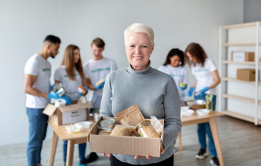 Fototapeta premium A happy senior woman holds a box filled with donation food, smiling at the camera. In the background, a group of volunteers is busy packing boxes for people in need, showing community spirit.
