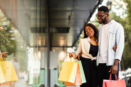 Seasonal Discounts. Portrair Of Smiling African Spouses Walking With Shopping Bags Outdoors, Lady Embracing Her Man And Pointing Finger Aside At Mall Window, Showing Product And Shopper Deals