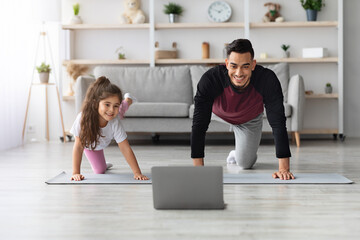 Obraz premium Happy arab father and little daughter having online fitness class, exercising at home together, wearing sportswear, training on yoga mat in front of laptop, enjoying workout, copy space
