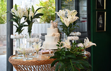 Elegant wedding dessert table with white tiered cake and lily floral arrangements, sophisticated reception decor with sweets display and religious icon, bright event venue interior setting