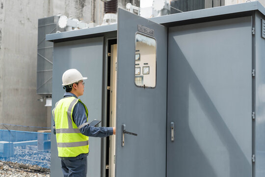 Power substation engineer opening electrical control cabinet during routine field inspection, recording measurements on tablet to ensure equipment safety, system reliability, and utility operations