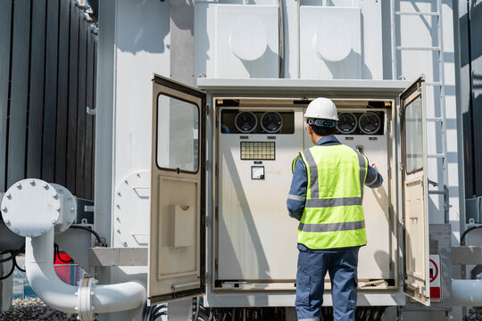 Professional electrical engineer inspecting control panel and monitoring gauges of power transformer at high voltage substation to ensure energy infrastructure efficiency and safety.