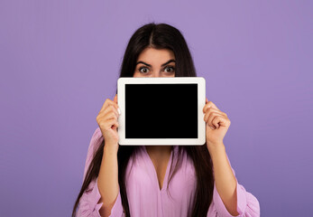 Naklejka premium Pretty armenian woman hiding her face behind digital tablet with blank screen, lady showing nice dating mobile application on pad, mockup, violet studio background
