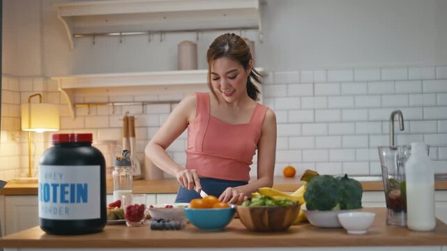 Smiling young Asian woman in pink top preparing a protein shake in modern kitchen,standing of protein powder and fresh fruits,vegetables.Concept of healthy meal prep,fitness nutrition and wellness