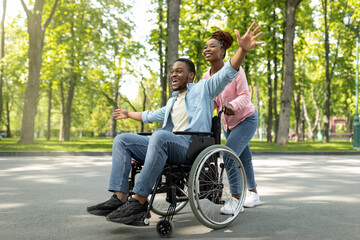 Happy black disabled guy in wheelchair on walk with his loving girlfriend outdoors, having fun, spending time together. African American impaired couple enjoying each other's company at green park