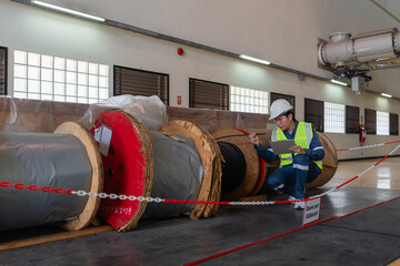 Engineer documenting large industrial cable reels stored inside a facility, verifying spare parts condition, inventory records, safety zones, and maintenance readiness for power infrastructure assets.