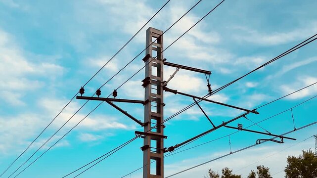 Utility pole against blue sky, birds perched on wires, concrete pylon and insulators under wispy clouds, suburban treeline below, calm atmospheric light, electrical infrastructure