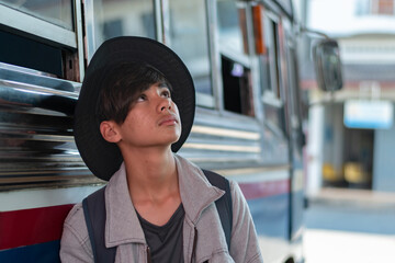 Pensive young male traveler with a backpack and hat, looking up while standing next to a bus. Focus on the feeling of anticipation, urban travel, and a sense of journey.