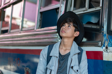 Pensive young male traveler with a backpack and hat, looking up while standing next to a bus. Focus on the feeling of anticipation, urban travel, and a sense of journey.