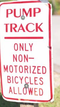 Verti al screen angled pump track sign Johnson City park near trailhead, bold red lettering on white metal plate declaring only non-motorized bicycles allowed, sunny neighborhood setting,