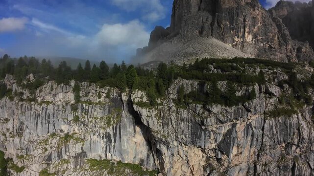 dramatic vertical cliff with pine trees, sweeping Dolomites panorama; hiker viewpoint captures rugged rock face, sunlight casting deep shadows, textured limestone cliffs, dense forest below,
