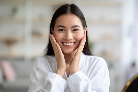 Closeup of asian woman in white bathrobe touching her skin and smiling, enjoying her young smooth skin, copy space. Cheerful young chinese lady touching her cheeks with both hands, home interior