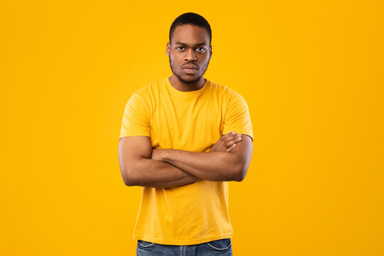 Serious African American Man Posing Crossing Hands Looking At Camera Frowning Expressing Negative Emotions Standing Over Yellow Background. Studio Shot. Male Emotional Blockage Problem - Powered by Adobe