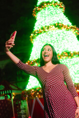Latina woman taking holiday selfie by illuminated christmas tree