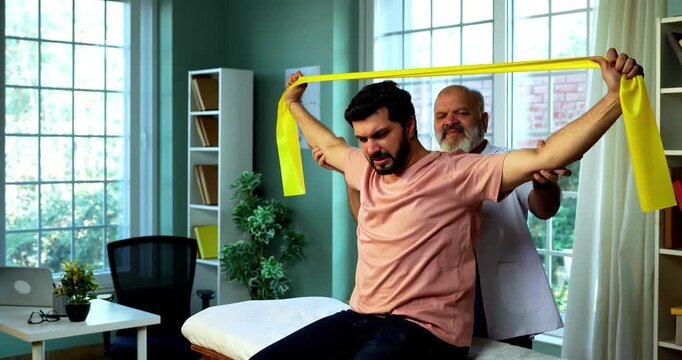 Indian physiotherapist teaching patient resistance band exercises for shoulder and tricep as senior male therapist assists strength training on clinic bed inside modern Indian clinic