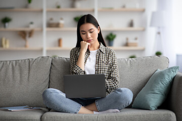Pensive young asian lady sitting on couch and using laptop, looking for job online from home, copy space. Chinese woman student studying, reading articles on Internet, using notebook