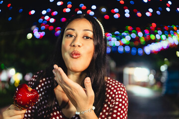 Young hispanic woman blowing kiss holding candied apple at night