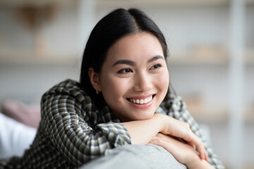 Closeup portrait of happy young asian lady enjoying weekend at home, smiling and looking at copy space. Cheerful korean woman reclining on couch alone in living room, resting