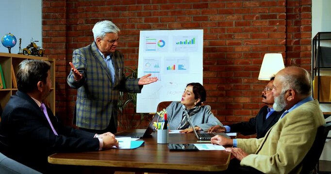 Indian senior business people arguing in modern office as group of senior Indian Asian businessmen engage in heated discussion over company growth, strategy while working with laptop, papers at desk