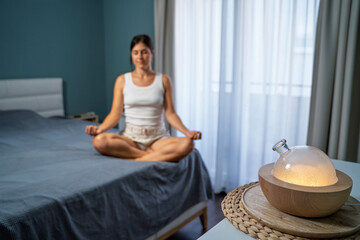 Young woman meditating in her bedroom while air diffusor is running, focus on diffusor