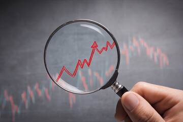 A hand holds a magnifier over a rising red arrow and chart on a dark background