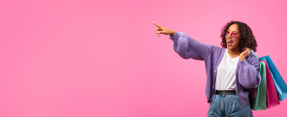 A young woman stands in front of a pink wall, wearing casual clothes. She is smiling and pointing with one hand while holding shopping bags in the other. The scene captures the excitement of shopping.