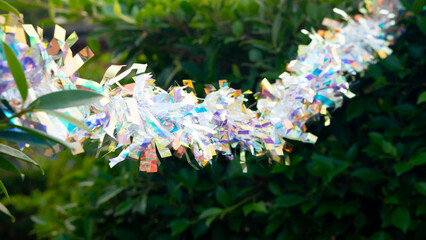 Colorful New Year festival decorations hanging on tree branches with green leaves background. Bright rainbow colors. Outdoor holiday celebration and seasonal greeting concept.