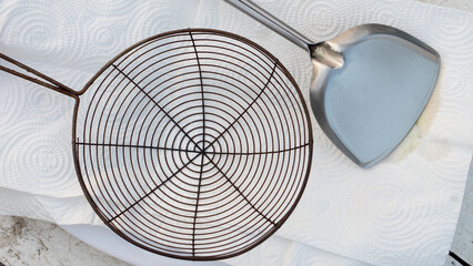 Top view of wire mesh strainer and stainless steel spatula on white paper towel. Kitchen utensils for frying and draining oil concept.
