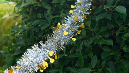 Colorful New Year festival decorations hanging on tree branches with green leaves background. Silver mixed with gold. Outdoor holiday celebration and seasonal greeting concept.