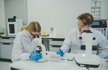 Female scientist examining specimen with microscope in laboratory while male colleague takes notes supporting teamwork in medical research