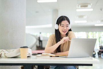 Glasses asian woman college student holding pen looking at laptop while sitting at table in building