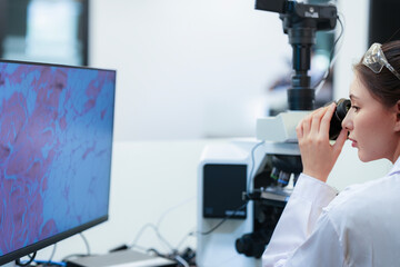 Young female scientist looking through microscope next to monitor with histology image. Researcher...
