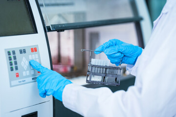 Medical researcher operating digital analyzer interface in sterile laboratory. Specialist holding biological samples in rack while setting calibration for pharmaceutical testing.