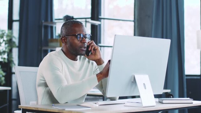 A focused businessman is talking on the phone while working on a computer in a contemporary office. Bright natural light fills the space, creating a productive atmosphere.
