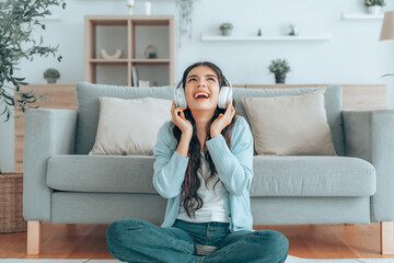 Young asian woman listening to music on couch in living room at home. Happy asia female using mobile smartphone, wearing headset and sitting on sofa