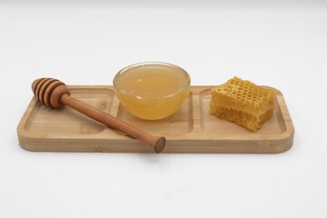 Glass bowl filled with honey, honeycomb piece and wooden dipper on bamboo tray. Clean white background, natural sweet food and organic ingredient concept.