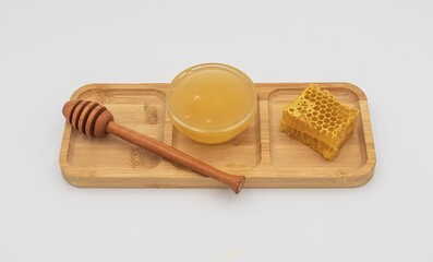 Glass bowl filled with honey, honeycomb piece and wooden dipper on bamboo tray. Clean white background, natural sweet food and organic ingredient concept.