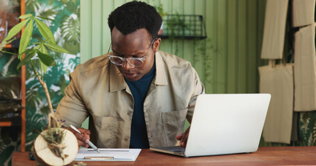 Laptop, documents and plant with black man in store for delivery checklist, account report and botany review. Order confirmation, ecommerce and florist entrepreneur with person in small business