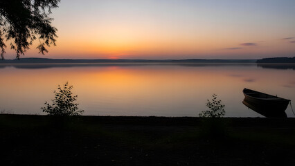 Calm lake water reflecting a golden sunset with a small wooden boat moored near the shore