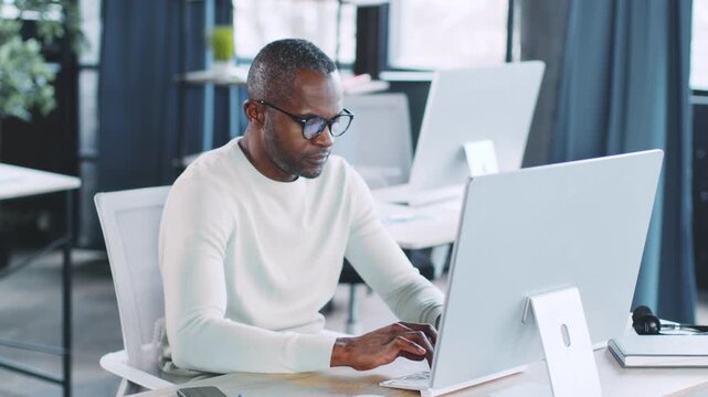 A man focused on his work sits at a sleek desk in a contemporary office. Natural light streams through large windows, enhancing the productivity and ambiance. He types attentively on a computer.