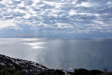 Fototapeta premium Wide-angle view from a Paphos cliffside, Cyprus. Dramatic sunbeams pierce heavy clouds over the shimmering Mediterranean Sea. Coastal landscape with rocky foreground and ethereal light.