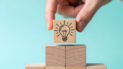 A hand placing a wooden block with a lightbulb idea on top of stacked blocks