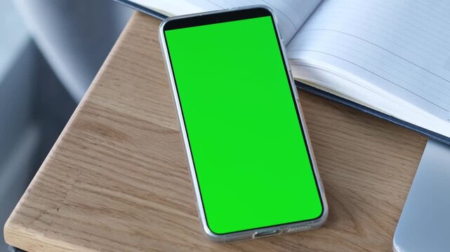 A modern cell phone with a chroma key green screen mockup lying flat on a sleek wooden table