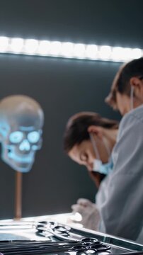 Forensic scientists wearing lab coats and masks are examining a body in a futuristic morgue, with a glowing skull model in the background, showing multiple shots of the same scene